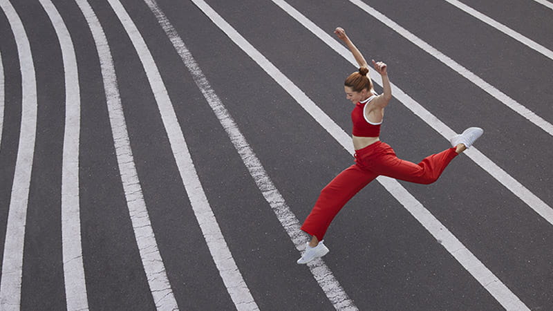 A woman in red athletic apparel is leaping over white painted lines on an asphalt surface. A woman in red athletic apparel is leaping over white painted lines on an asphalt surface.