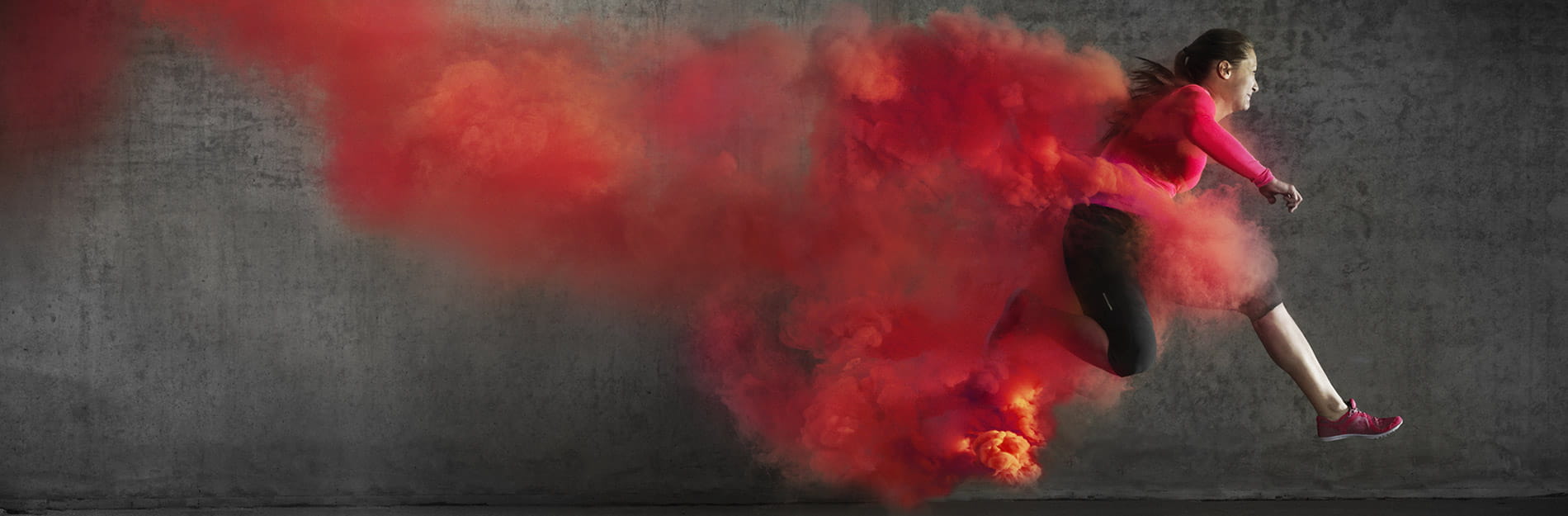 A woman in athletic apparel is running through a trail of red smoke on a grey background. A woman in athletic apparel is running through a trail of red smoke on a grey background.