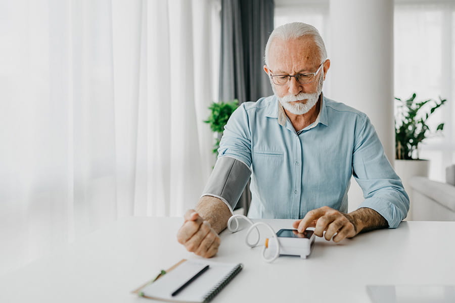 older man at home at a table, checking his blood pressure with a cuff and monitor kit